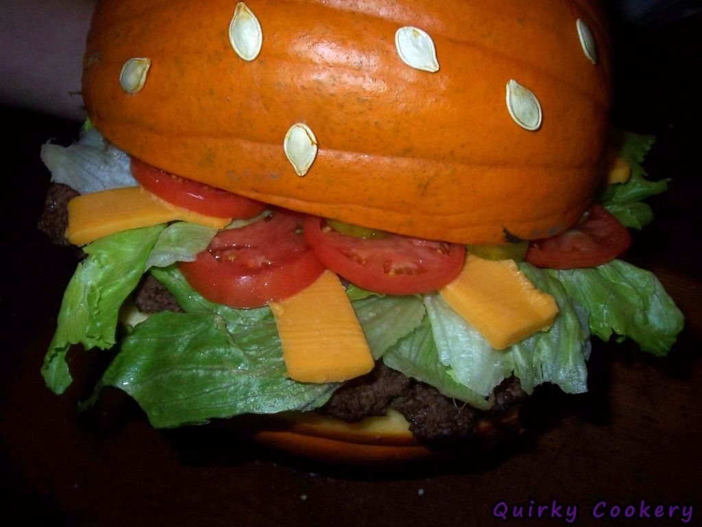 Giant fake burger made out of a whole pumpkin to look like the bun with a large burger patty and toppings inside. Pumpkin seeds glued to the outside to look like a sesame seed bun. 