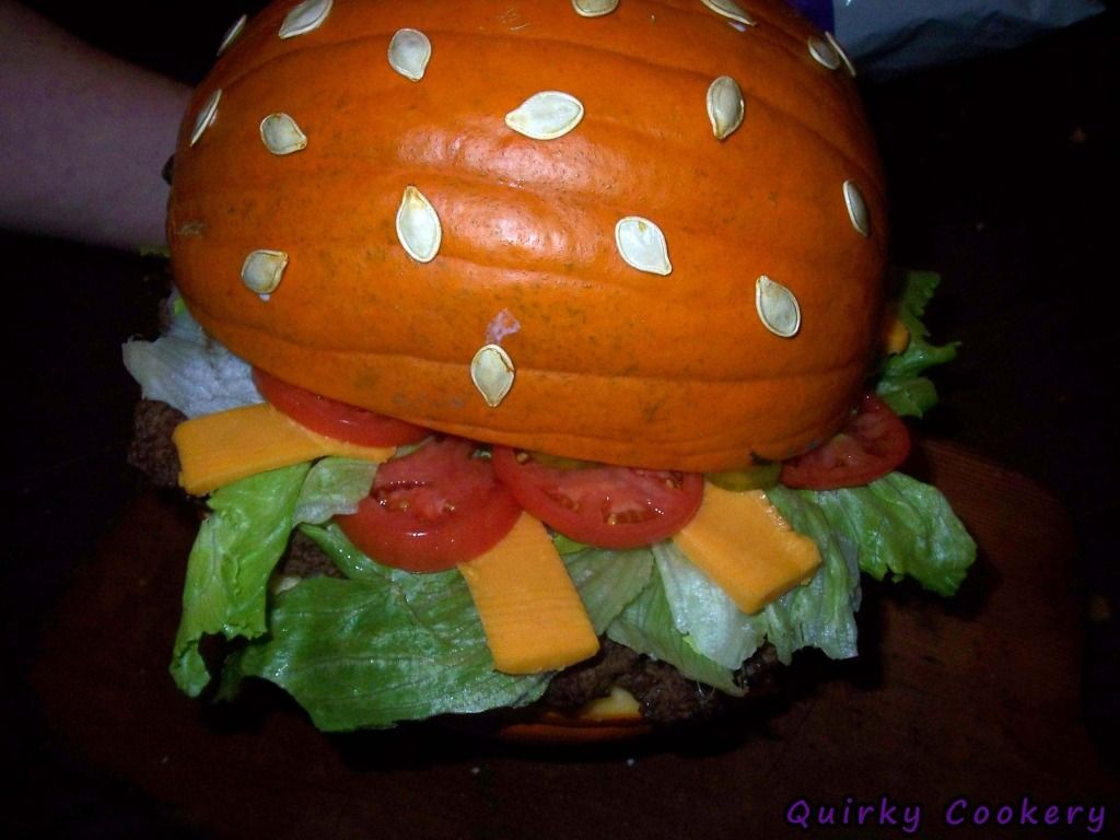 Giant fake burger made out of a whole pumpkin to look like the bun with a large burger patty and toppings inside. Pumpkin seeds glued to the outside to look like a sesame seed bun. 