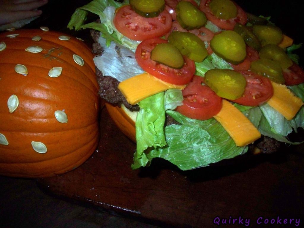 Giant fake burger made out of a whole pumpkin to look like the bun with a large burger patty and toppings inside. Pumpkin seeds glued to the outside to look like a sesame seed bun. 